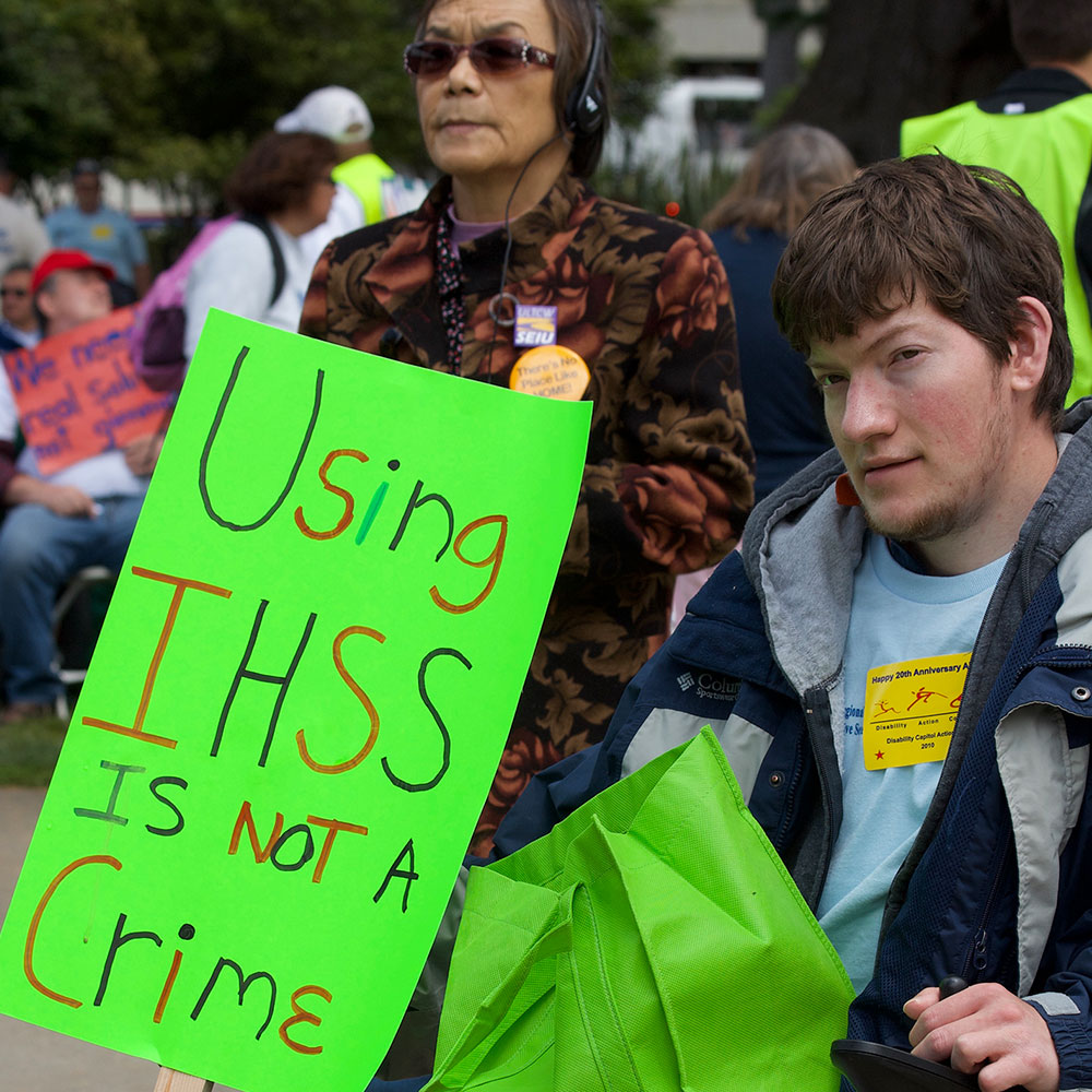 Photo of man at advocacy rally with a sign saying, "Using IHSS is not a crime."