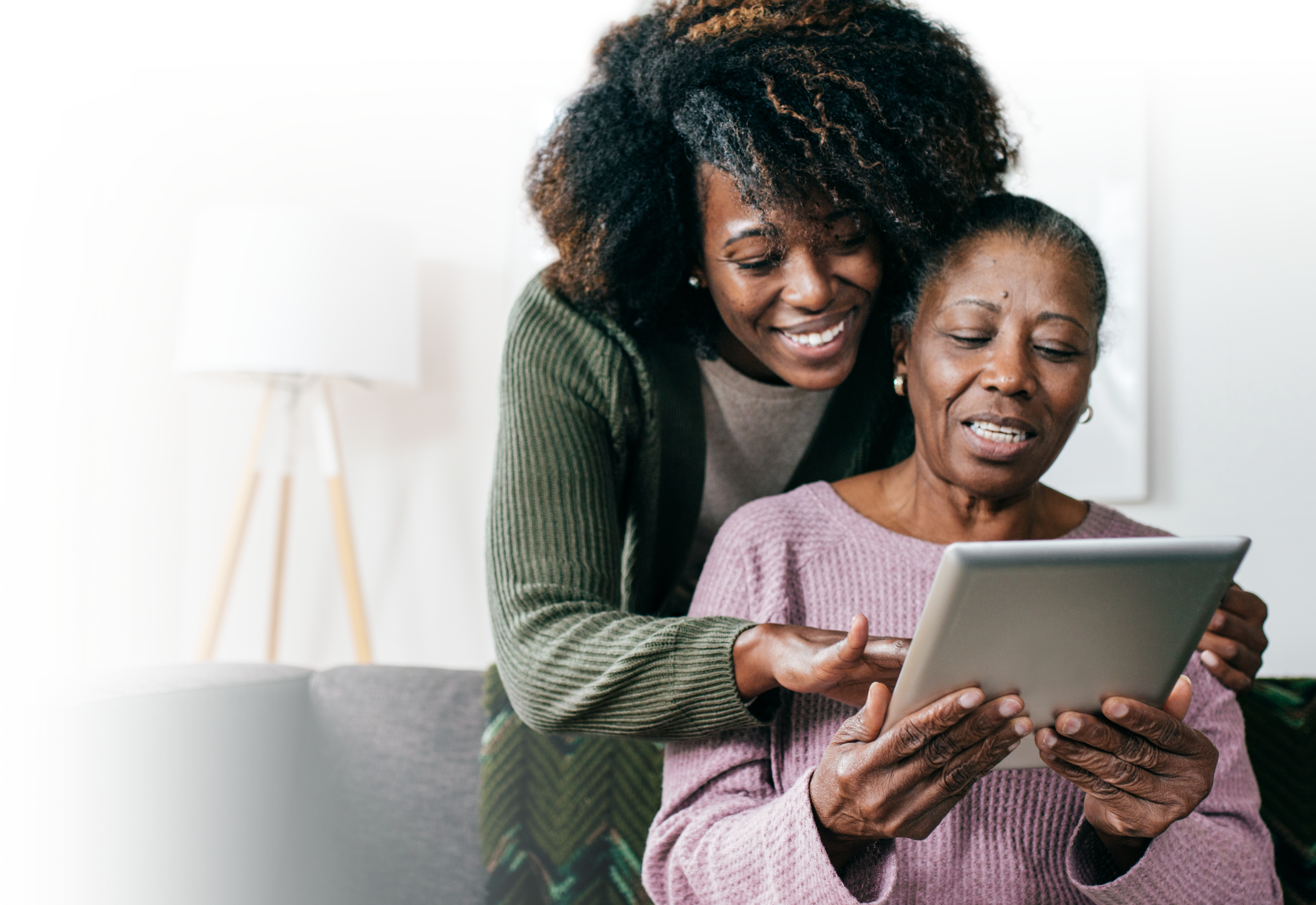 Photo of senior woman on the couch with her adult daughter helping her use a digital tablet.